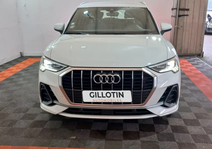 Audi Q3 blanc vue de face, avec calandre large à barres verticales et phares LED allumés dans un garage.