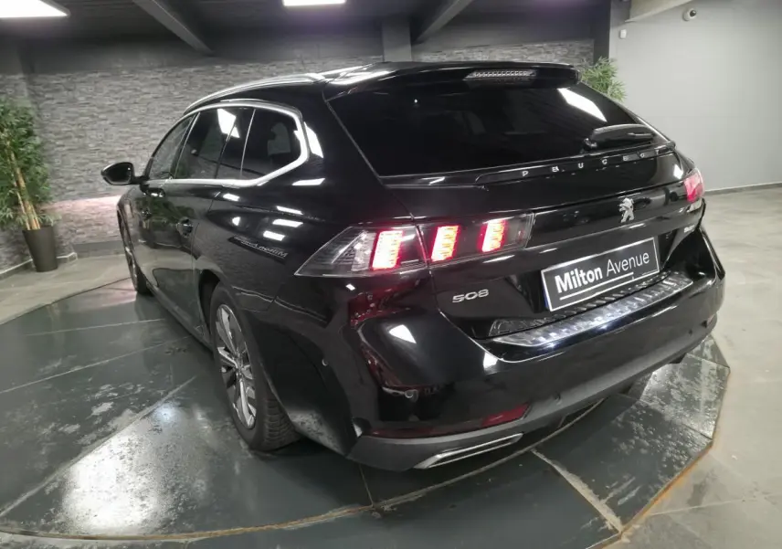 Peugeot 508 SW noire vue 3/4 arrière droit, feux arrière allumés et barres de toit visibles.