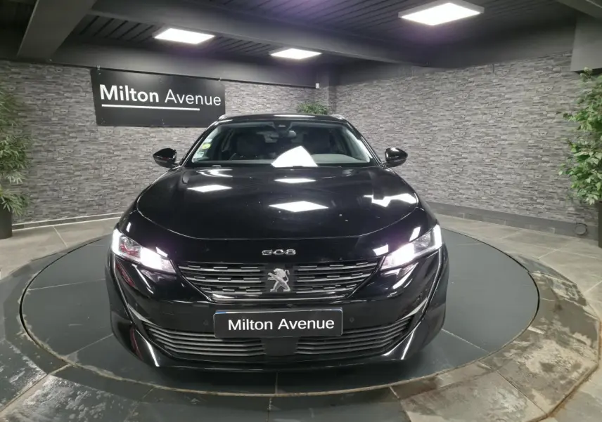 Peugeot 508 SW noire vue de face dans un showroom, avec calandre chromée et phares allumés.