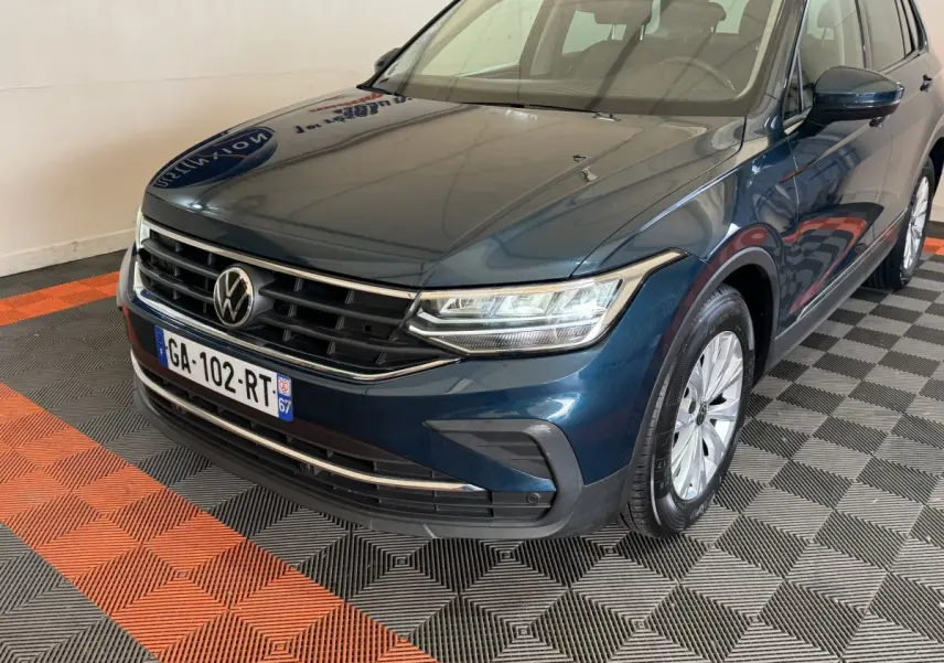 Volkswagen Tiguan bleu en 3/4 avant droit, avec calandre chromée et phares LED allumés, sur sol damier noir et orange.