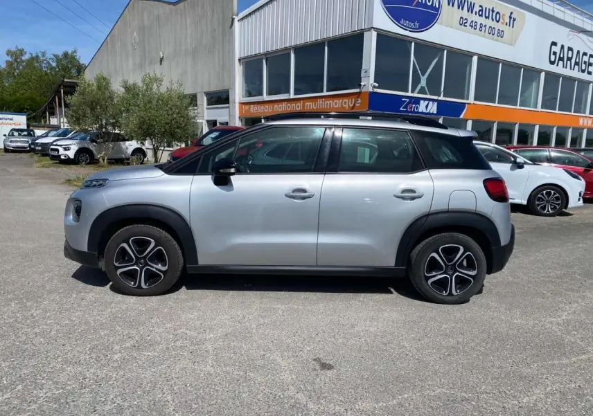 Vue de profil côté gauche d'un Citroën C3 Aircross gris Artense avec toit noir et jantes distinctives, stationné devant un garage.