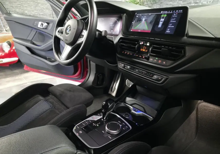 Vue intérieure côté conducteur d'une BMW Série 1 116d rouge, avec console centrale moderne et écran tactile affichant la caméra de recul.