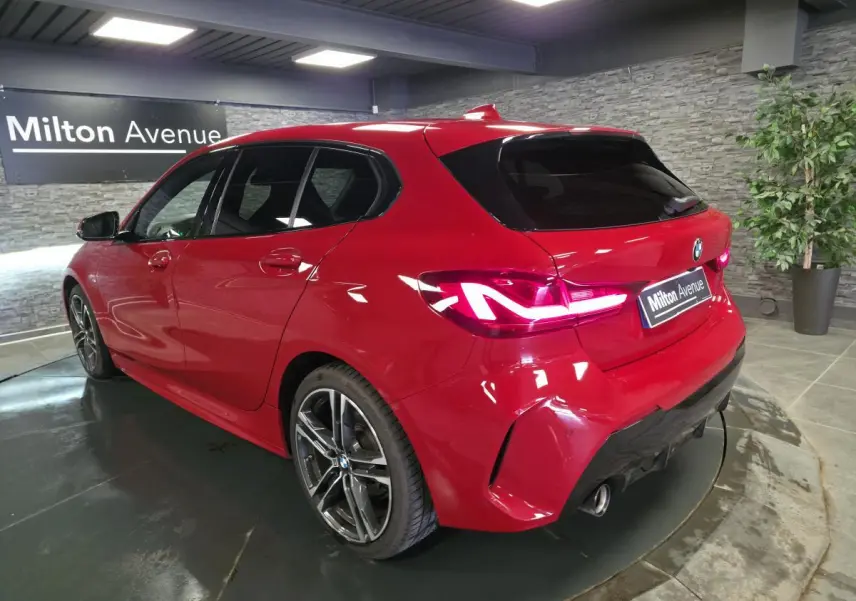 BMW Série 1 116d M Sport rouge vue 3/4 arrière droit, avec jantes alliage 18 pouces et feux LED allumés.