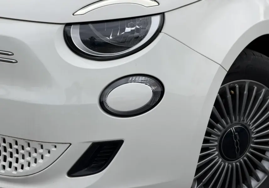 Gros plan sur l'avant droit blanc d'une FIAT 500 électrique 2023, mettant en valeur le phare LED et la jante stylisée.