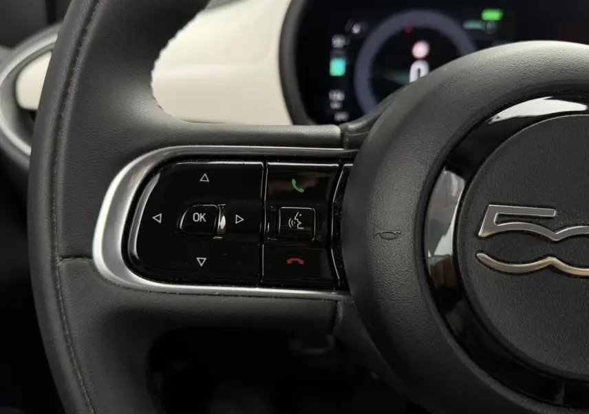 Gros plan sur le volant noir de la FIAT 500 électrique 2023 avec commandes intégrées et tableau de bord partiellement visible.