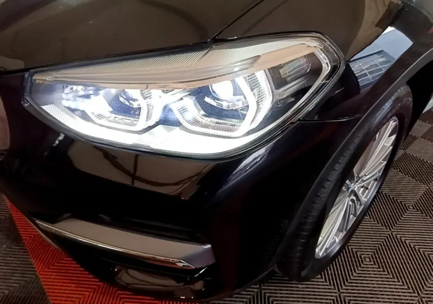 Gros plan sur le phare avant droit allumé d’un BMW X3 noir, avec jante alliage visible et carrosserie brillante.
