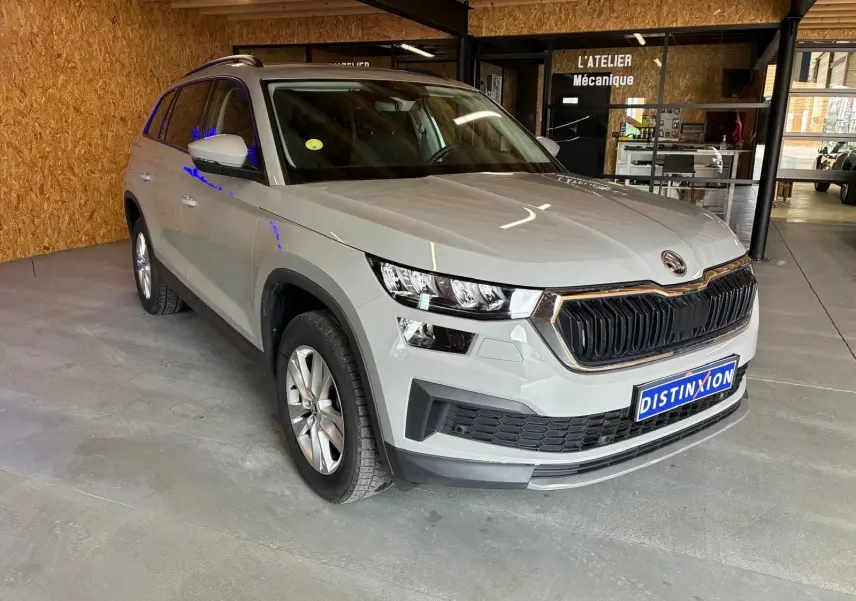 SKODA KODIAQ gris clair en 3/4 avant droit, SUV 7 places avec calandre noire et jantes argentées, stationné en intérieur.