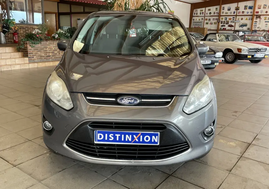 Vue frontale d'une Ford C-MAX gris métallisé 2012 avec calandre noire et plaque d'immatriculation Distinxion dans un showroom.