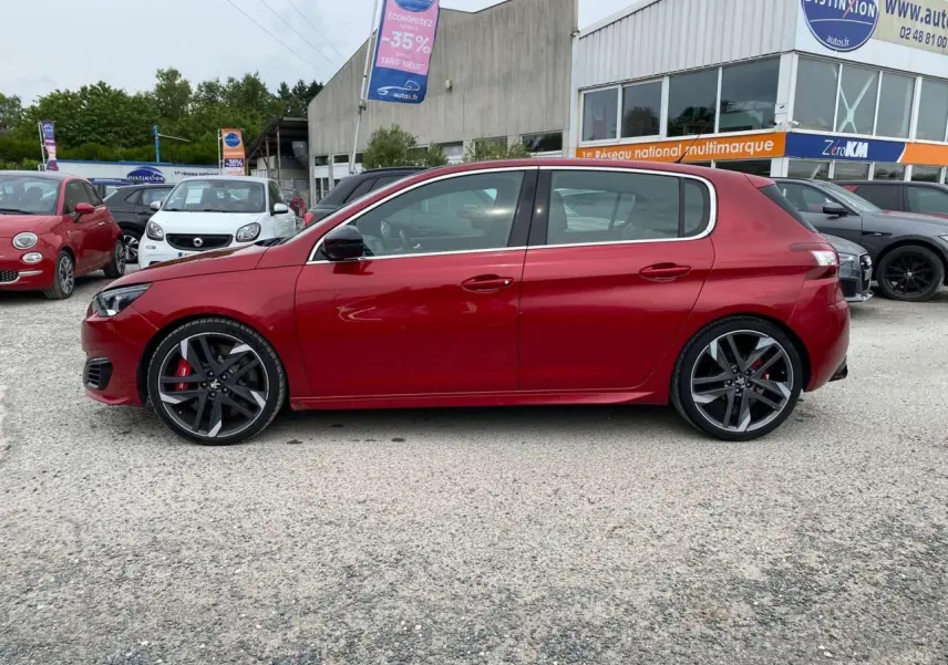 Profil latéral droit d'une Peugeot 308 GTi rouge métallisée garée devant un concessionnaire multimarque.