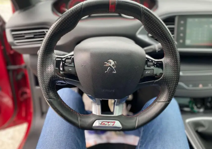 Volant cuir perforé avec logo GTi au centre d'une Peugeot 308 rouge, vue intérieure depuis le siège conducteur.
