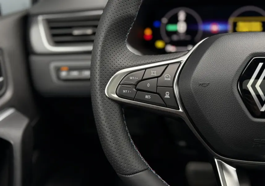 Gros plan sur le volant perforé avec commandes intégrées et logo Renault, tableau de bord numérique flou en arrière-plan.