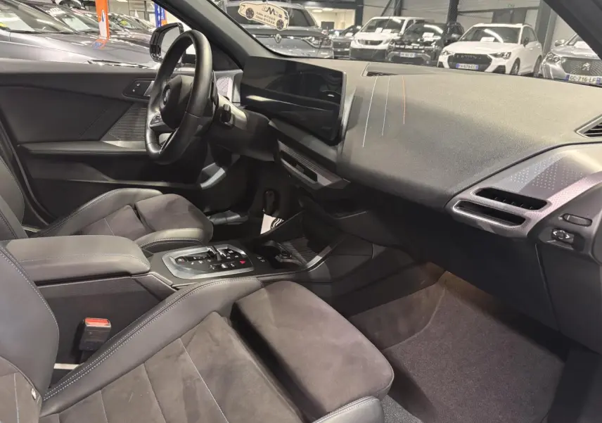 Intérieur BMW Série 1 118D 2025 vu côté conducteur, sellerie Alcantara noire avec surpiqûres bleues et tableau de bord moderne.