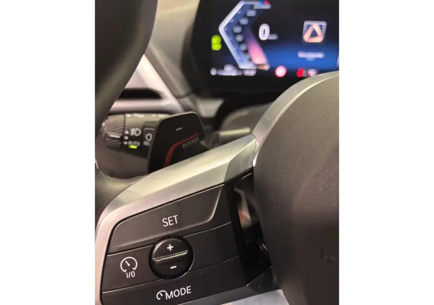 Gros plan sur les commandes du volant à gauche de la BMW Série 1 118D blanche, avec affichage numérique en arrière-plan.