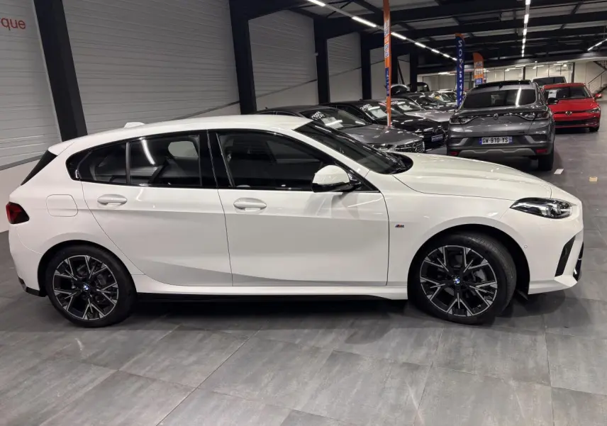 Vue de profil côté gauche d'une BMW Série 1 blanche avec jantes 18 pouces et finitions M Sport en intérieur showroom.