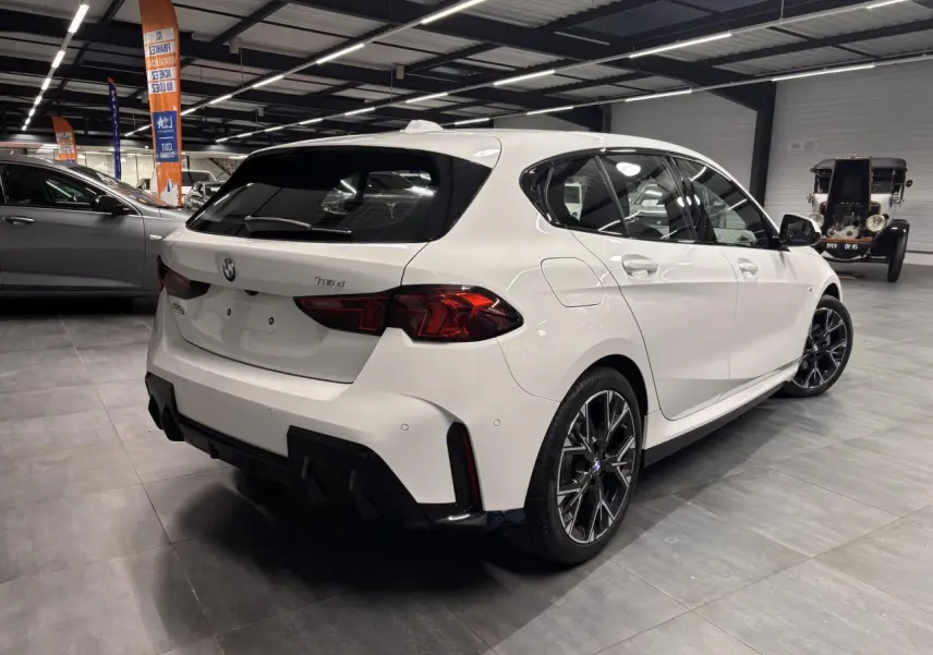 BMW Série 1 118D blanche vue 3/4 arrière droit en intérieur, avec jantes 18 pouces et éléments M Sport noirs.