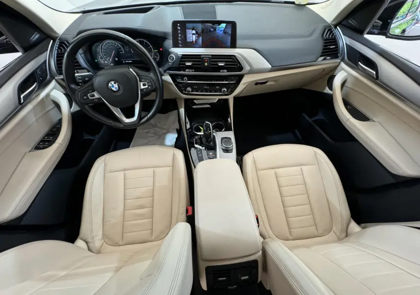 Vue intérieure avant de la BMW X3 2018 avec sièges cuir beige, tableau de bord noir et écran tactile central allumé.
