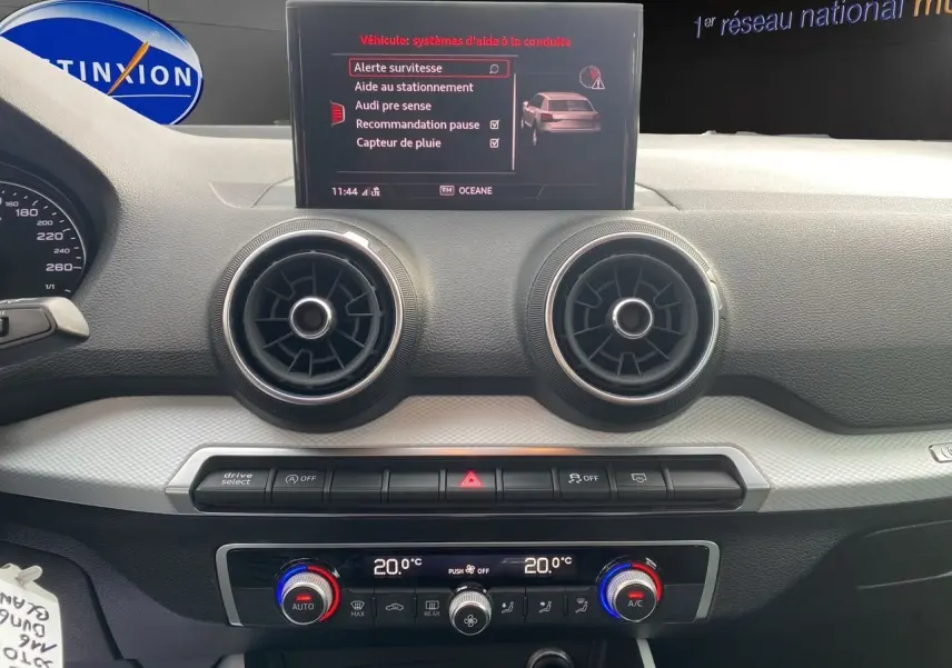 Tableau de bord de l'Audi Q2 blanc 2019, affichage central avec menu d'aides à la conduite et commandes climatisation double zone.