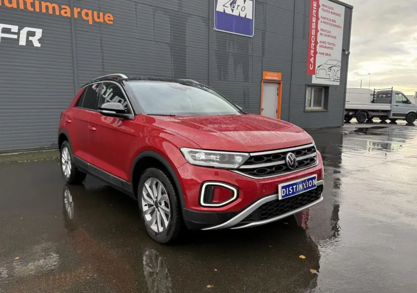 Volkswagen T-Roc rouge Kings Red métallisé vu en 3/4 avant droit sur sol mouillé devant un bâtiment gris.