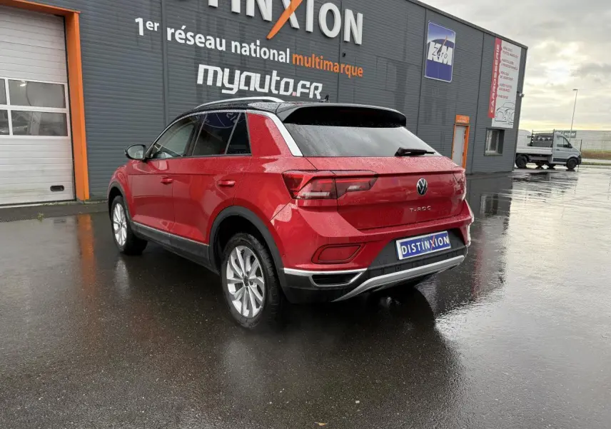 Volkswagen T-Roc rouge Kings Red métallisé vu en 3/4 arrière droit sur sol humide devant un bâtiment gris.