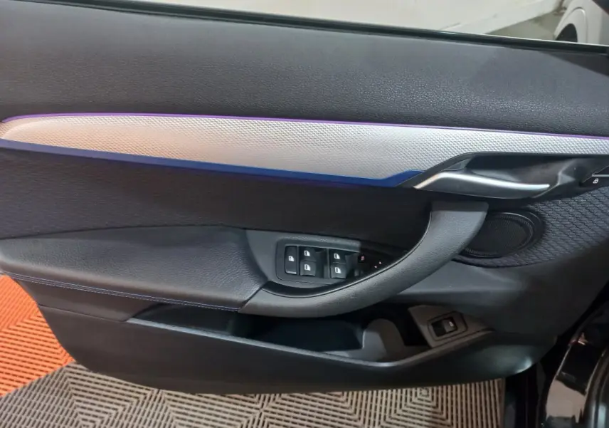 Vue intérieure côté gauche de la porte noire du BMW X1 xDrive20d M Sport avec éclairage d'ambiance bleu et commandes fenêtres.