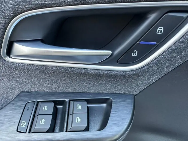 Gros plan sur la poignée intérieure côté conducteur et les commandes de vitres d’une MG ZS Hybrid+ blanche, tissu noir.