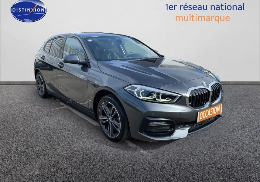 BMW Série 1 gris minéral métal vue 3/4 avant droit avec calandre noire et phares LED allumés.