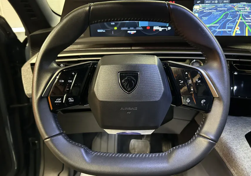 Vue rapprochée du volant noir du Peugeot 3008 III Hybrid 145 e-DSC6 Allure avec commandes intégrées et écran digital en arrière-plan.