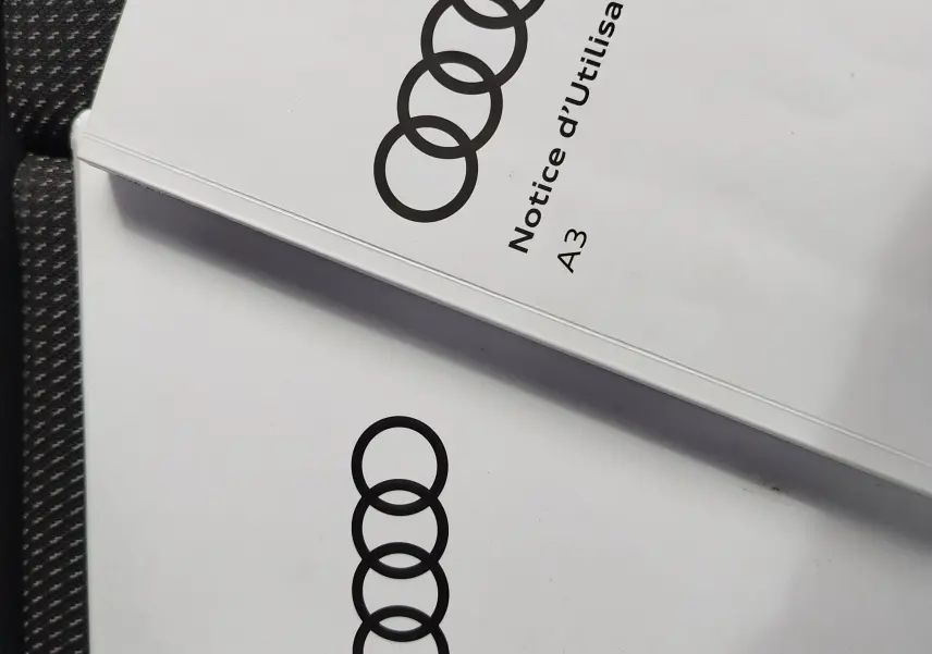 Image ne montrant pas le véhicule mais deux notices d'utilisation Audi A3 posées sur un siège en tissu gris foncé à motifs.