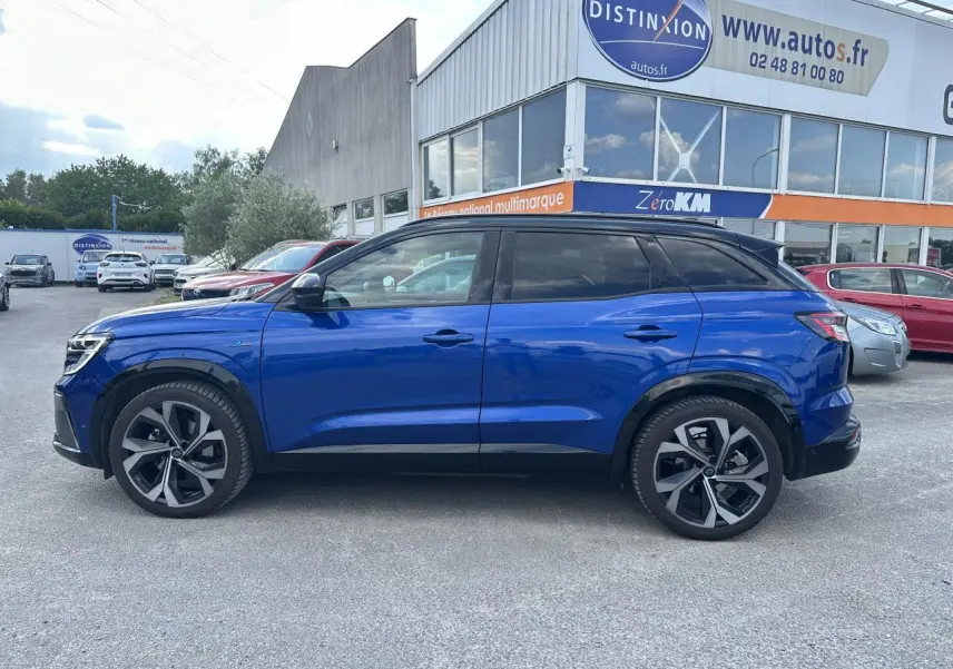 Profil droit du Renault Austral 2024 bleu avec toit noir, jantes alliage et vitres teintées, stationné devant un bâtiment.
