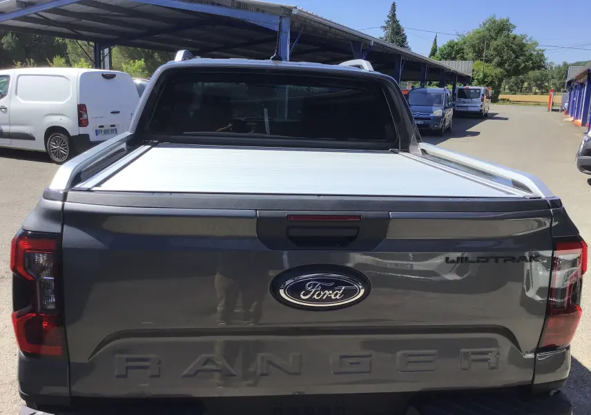 Vue arrière d'un Ford Ranger Double Cabine gris carbone avec rideau de plateau coulissante électrique et logo Wildtrak visible.