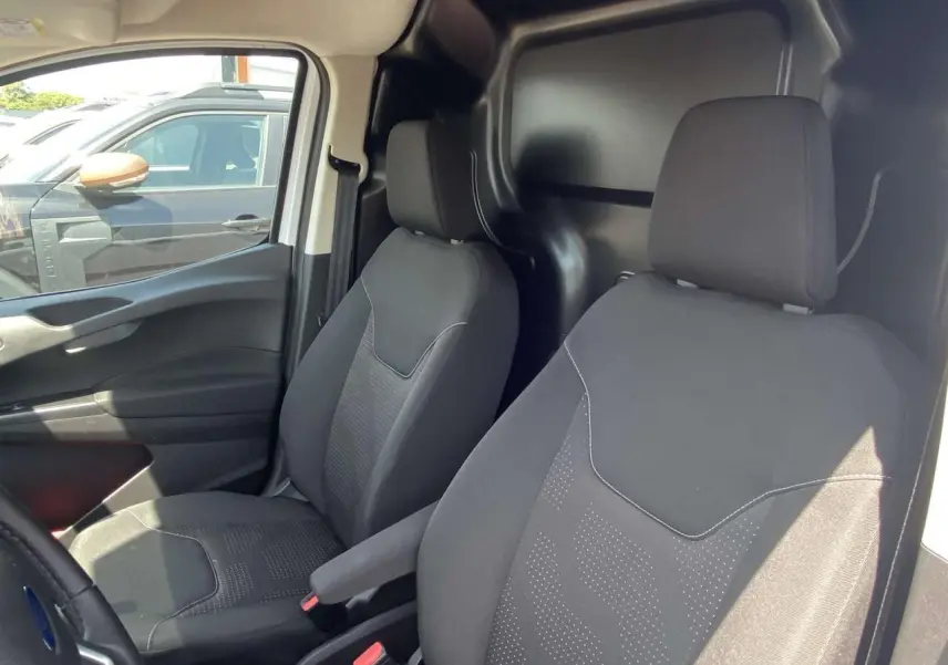 Vue intérieure côté conducteur et passager du Ford Tourneo Courier blanc, montrant les sièges noirs et la cloison tôlée arrière.