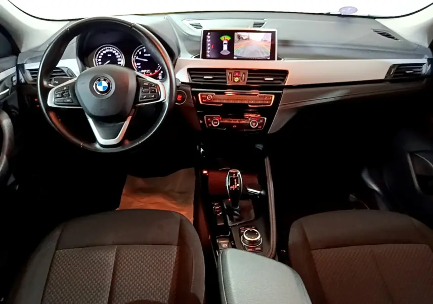 Vue intérieure avant du BMW X2 sDrive18i Lounge 2023, volant cuir alu multifonctions et écran central affichant l'aide au stationnement.