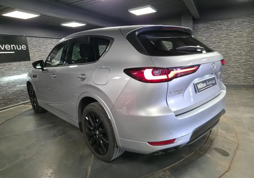 Vue 3/4 arrière droite du Mazda CX-60 gris Sonic Silver avec jantes noires et feux arrière LED allumés en intérieur showroom.