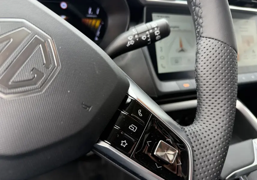 Gros plan sur le volant perforé noir du MG MOTOR ZS 1.5 HYBRID+ 197 LUXURY avec commandes intégrées et tableau de bord numérique flou.