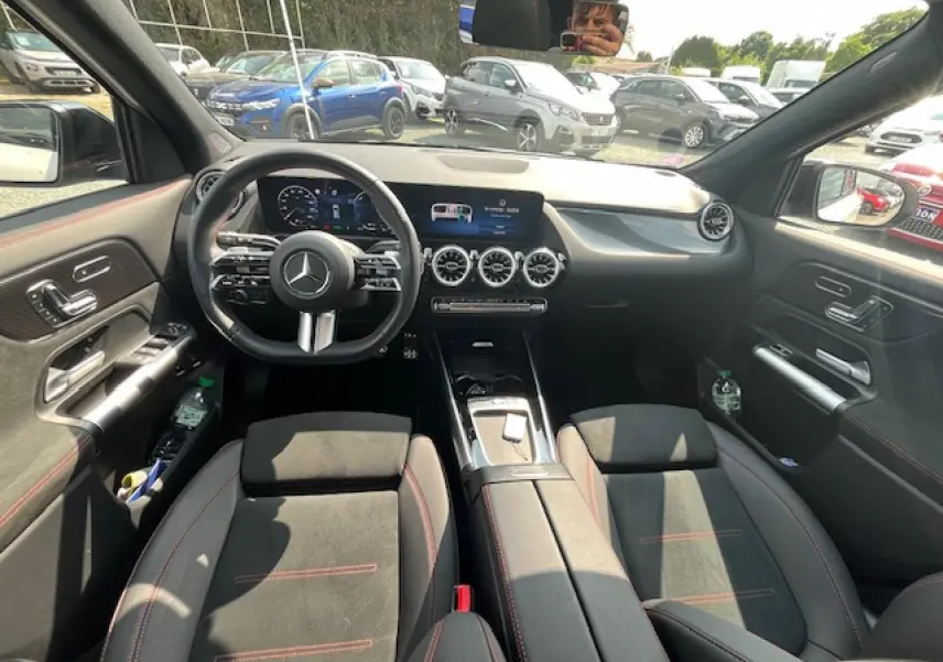 Intérieur du Mercedes GLA 250 e hybride 2024, vue avant du poste de conduite avec écran central et sièges noirs à surpiqûres rouges.