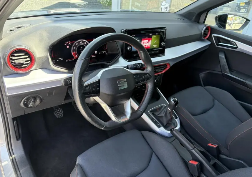 Intérieur de la SEAT Arona 2024 vu côté conducteur, volant sport cuir noir avec surpiqûres rouges et tableau de bord digital.