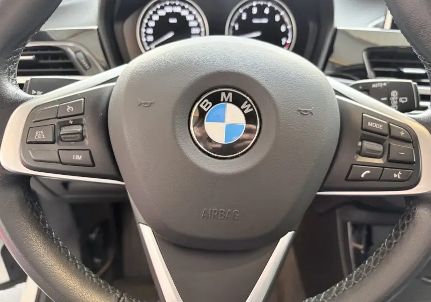 Vue rapprochée du volant cuir et alu multifonctions d'une BMW X1 blanche avec tableau de bord numérique en arrière-plan.