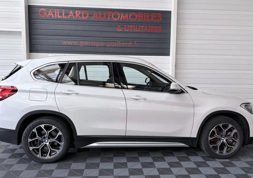 Profil droit d’un BMW X1 blanc 2021 avec jantes alliage et rails de toit chromés, stationné en intérieur.