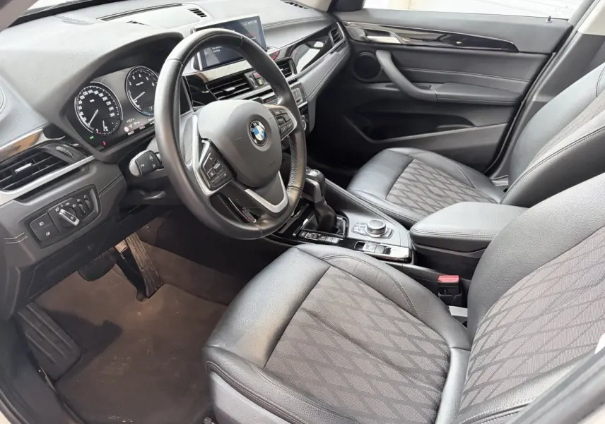 Vue intérieure côté conducteur d'une BMW X1 xDrive25eA xLine 2021 avec volant cuir alu multifonctions et sièges tissu noirs.