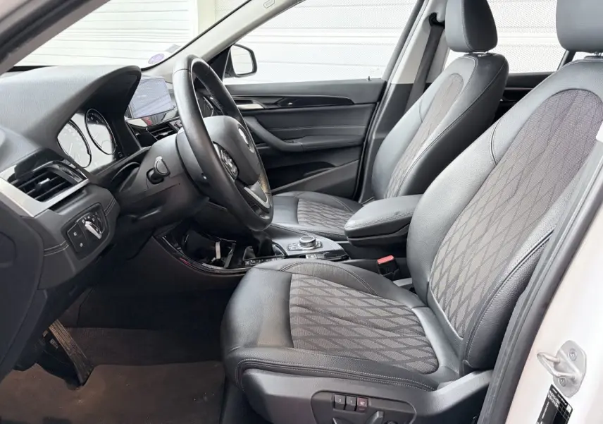 Vue intérieure côté conducteur de la BMW X1 blanche, montrant sièges en cuir et tissu avec motifs géométriques.
