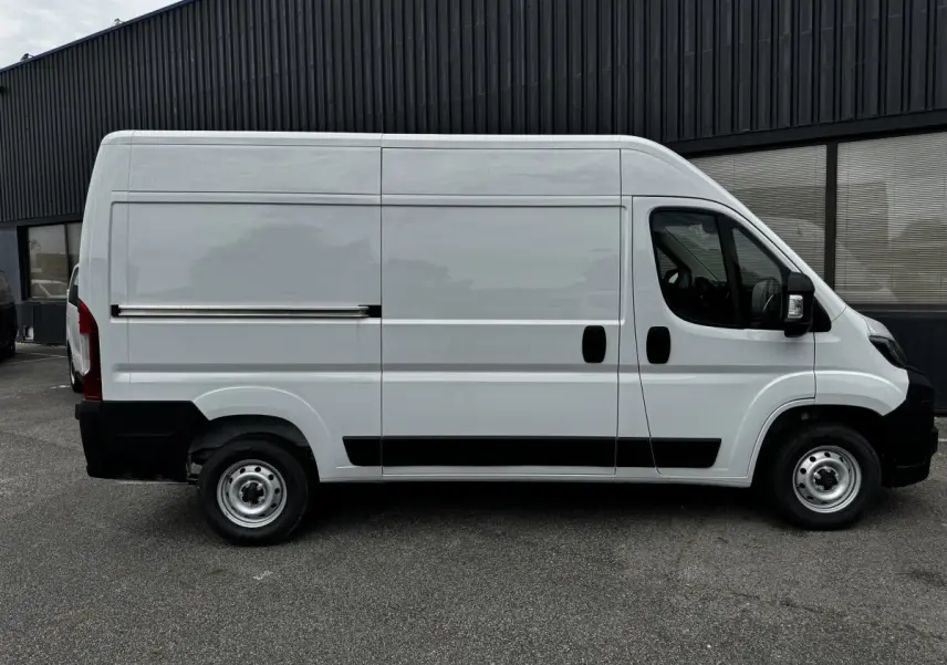 Vue de profil côté droit d'un fourgon Peugeot Boxer blanc avec porte coulissante et protections noires sur un sol goudronné.