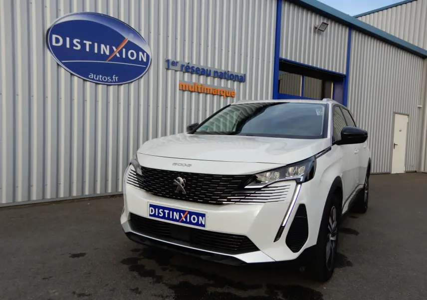 Peugeot 5008 blanc nacré en 3/4 avant droit, avec calandre noire et feux LED distinctifs, devant un bâtiment industriel.