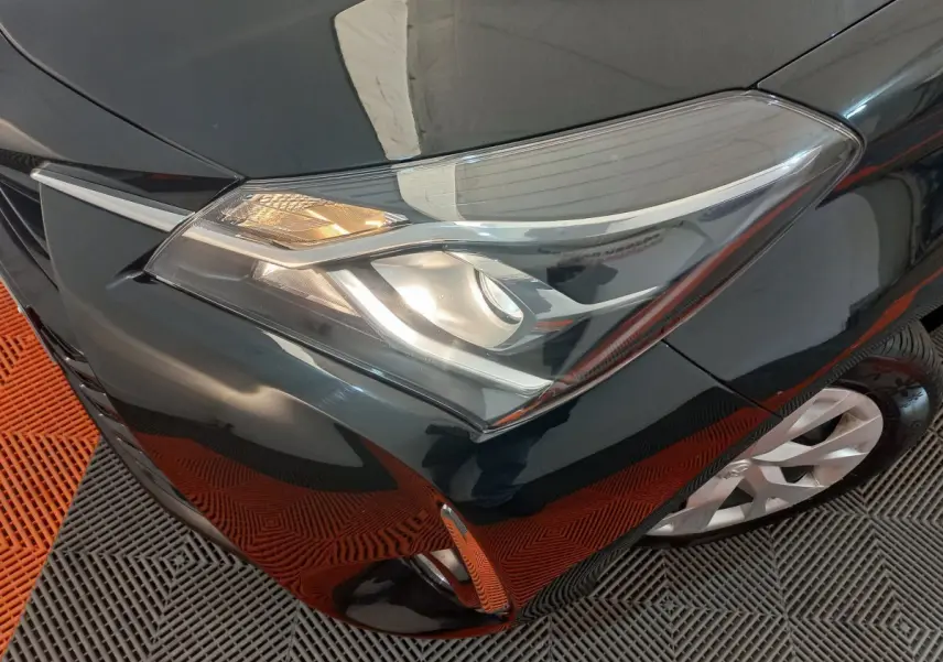 Gros plan sur le phare avant gauche d'une Toyota Yaris noire, avec partie de la roue et pare-chocs visibles.