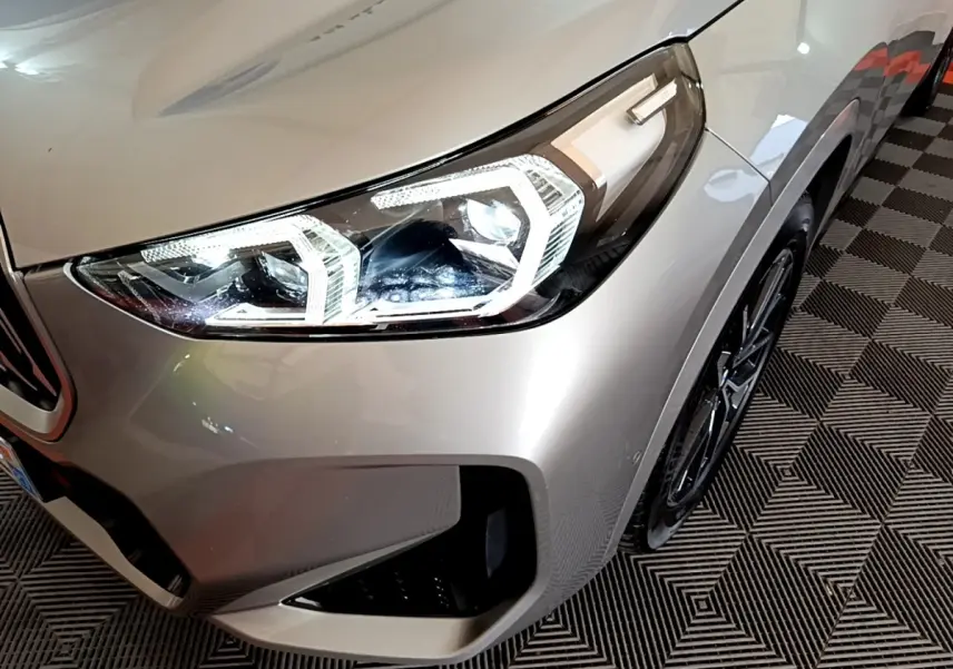 Gros plan sur l'avant droit gris clair du BMW X1 xDrive23i M Sport 2023, mettant en valeur le phare LED et la jante noire.