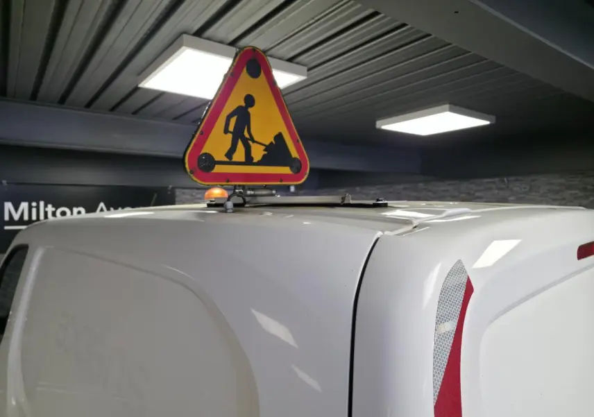 Vue arrière droite d’un Peugeot Partner blanc avec panneau de signalisation travaux fixé sur le toit en intérieur.