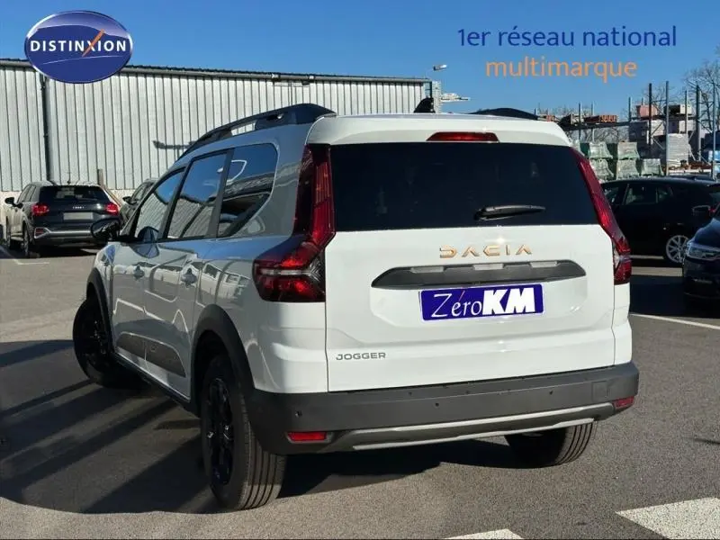 Vue 3/4 arrière droite d'un Dacia Jogger blanc Glacier avec barres de toit et jantes noires sur parking.