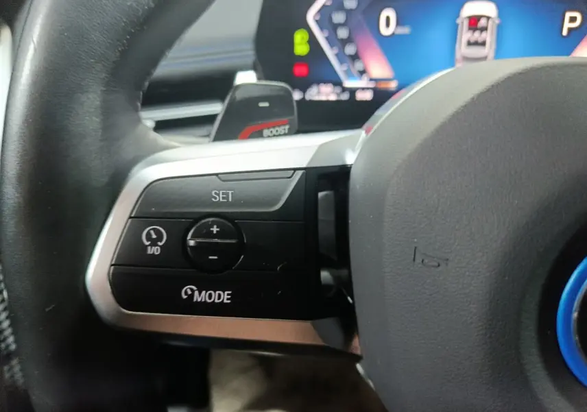 Gros plan sur les commandes du volant cuir noir du BMW X1 xDrive25e blanc, avec affichage numérique en arrière-plan.