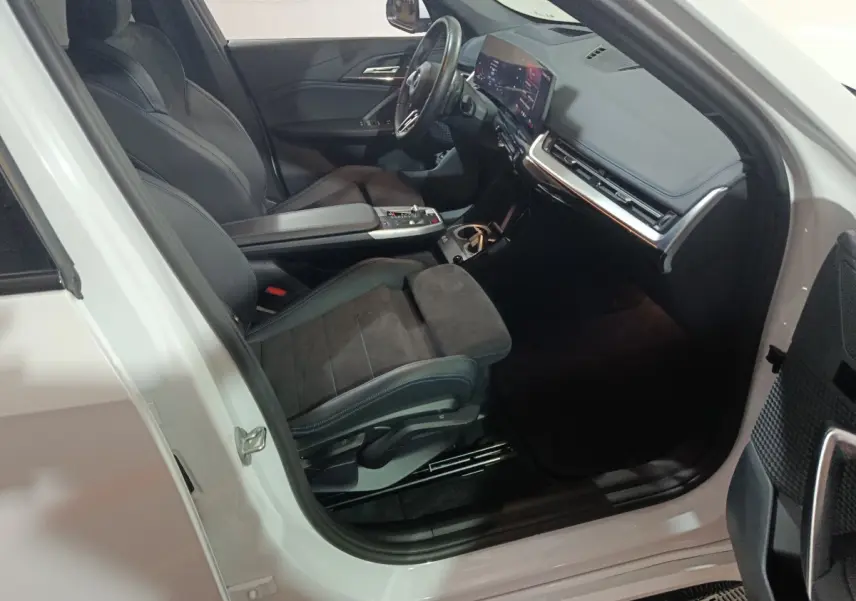 Vue intérieure côté conducteur d'une BMW X1 blanche 2024, avec sièges noirs et tableau de bord digital lumineux.