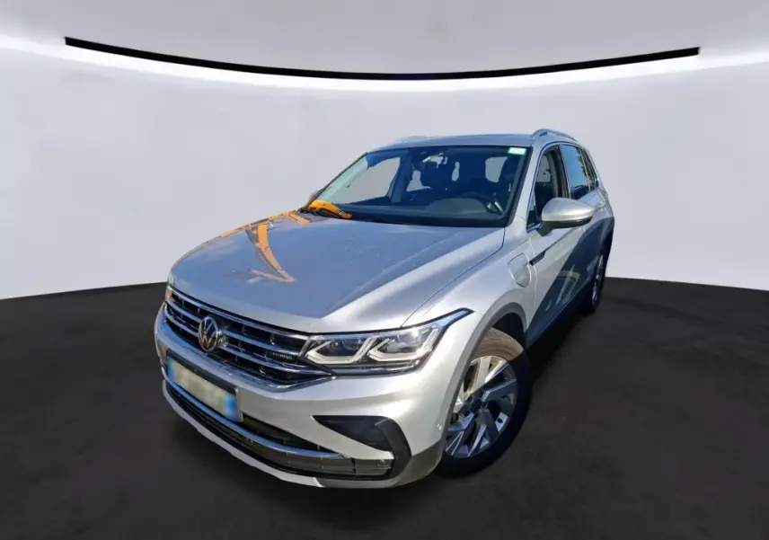Vue 3/4 avant d'un Volkswagen Tiguan gris clair hybride rechargeable, avec calandre chromée et phares LED visibles.