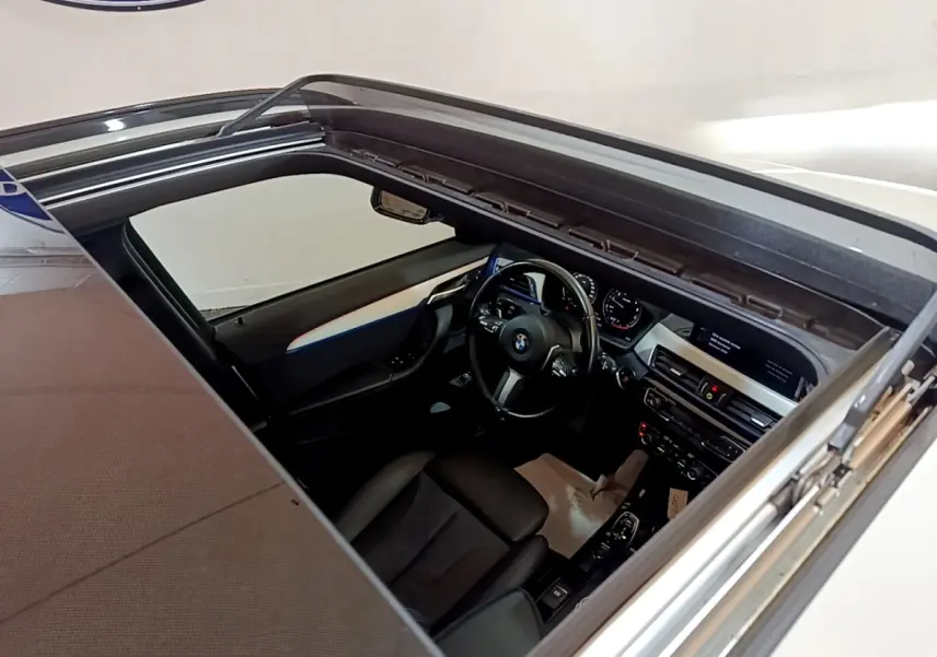 Vue intérieure par le toit ouvrant d'une BMW X1 blanc, montrant le tableau de bord et volant cuir multifonctions.